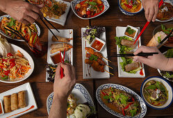 Verschiedene pikante thailändische Speisen auf Holztisch, Hände mit Essstäbchen