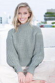 Junge blonde Frau in grauem Strickpullover und heller Hose am Strand