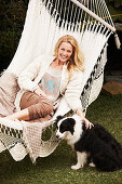 A blonde woman in a hammock wearing a light cardigan and brown trousers with a dog next to her