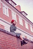 Junge Frau mit Hut in 60er Jahre Outfit auf Ziegelmauer sitzend