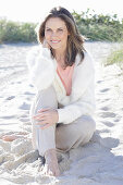 A long-haired woman sitting in the sand on the beach wearing a light jumper and trousers