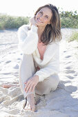 A long-haired woman sitting in the sand on the beach wearing a light jumper and trousers