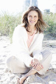 A long-haired woman sitting in the sand on the beach wearing a light jumper and trousers