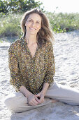 A long-haired woman sitting in the sand wearing a floral shirt and light trousers