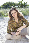 A long-haired woman sitting in the sand wearing a floral shirt and light trousers