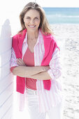 A long-haired woman on the beach wearing a long blouse with a pink jumper over her shoulders