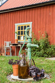 Brunnen, braune Flasche und Purpurglöckchen vorm roten Haus