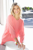 A young blonde woman wearing a salmon-pink blouse and white trousers