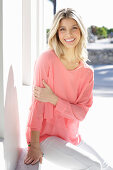 A young blonde woman wearing a salmon-pink blouse and white trousers