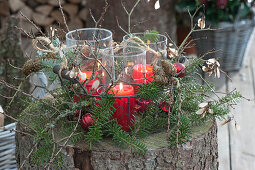 Windlichter im Drahtkorb, weihnachtlich dekoriert mit Zweigen von Tanne, Lärche, Ahorn und rotem Christbaumschmuck auf Baumstamm