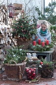 Christmas terrace with Christmas tree, Advent wreath, baskets with fir branches, pinecones and Christmas tree decorations, a mug with hot mulled cider on wooden disc, woman lighting candles, antlers, and old wooden skates as decoration