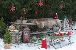 Bench with fur as a seat in front of spruce with red stars, tray with cups on a sled, basket with firewood, lanterns, cookie jar with cinnamon stars, thermos flasks