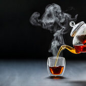 Dampfender schwarzer Tee wird aus Teekanne in Glasbecher gegossen