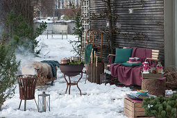 Terrasse für die Winterparty: Töpfe mit heißem Punsch auf dem Grill, Feuerkorb, Bank und Brennholzregal mit Fell, Decke, und Kissen als Sitzplätze, Stockbrot und Schlitten an Baumstamm