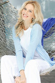 A blonde woman wearing a white top and a light blue cardigan sitting in a hammock