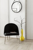 Branches in yellow vase next to black chair below mirror on wall