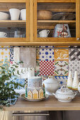 Christmas cake and soup tureens in front of colorful tiled backsplash, crockery cupboard above