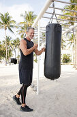 A grey-haired man wearing black sports clothing using a punching bag