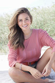 A young, long-haired woman wearing a pink blouse and shorts sitting in the sand