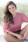 A young, long-haired woman wearing a pink blouse and shorts sitting in the sand