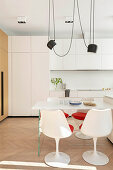 White dining table and white chairs with red seat cushions below pendant light with white fitted kitchen in background