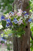 A small bouquet of columbines, wild strawberry and woodruff flowers in a glass jar with a grass wreath hung on a post