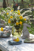 A set Easter table decoration in the garden: a bouquet of flowering twigs and dandelions, Easter bunny, basket with Easter eggs, and plates with napkins