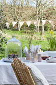 Table set for Easter meal outdoors with white grape hyacinths under glass cover below string of bunting