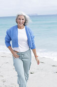 Reife Frau mit grauen Haaren in weißem T-Shirt, blauer Strickjacke und Hose am Strand