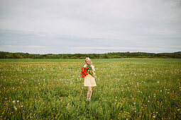 Junge blonde Frau im Kleid steht mit rotem Tulpenstrauß auf einer Wiese