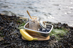 Overnight Oats mit Blaubeeren in Lunchbox und eine Banane am Ufer