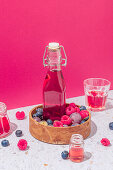 Glass bottle of fresh fruits juice in wooden bowl with ripe berries served on table with glasses on pink background