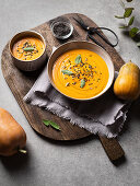 Pumpkin soup served with seasonings placed near fresh pumpkin on a wooden board on gray table