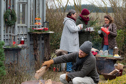 Man puts log in fire pit, in the background women with mulled wine