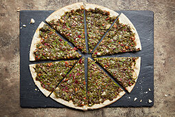 A pizza topped with leafy vegetables, pistachios, pine nuts and pomegranate, sliced on a slate platter