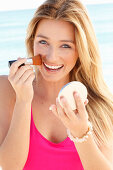 Young blonde woman in pink dress applying makeup with brush