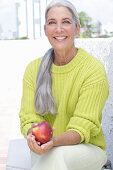A grey-haired woman wearing a greenish-yellow knitted jumper and light-coloured trousers holding an apple