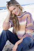 Young blond woman in knitted jumper with colour gradient and jeans by the sea