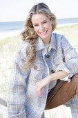 Young blonde woman in checked coat and suede trousers by the sea