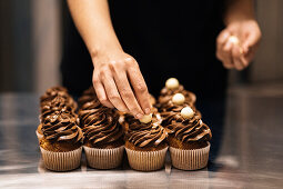 Unrecognizable woman putting sugar balls on top of yummy muffins with chocolate cream during work in bakehouse