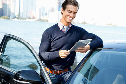 A young man wearing a jumper standing next to a car holding a tablet