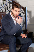 Young man with a beard in a jacket and jeans is sitting on a leather sofa