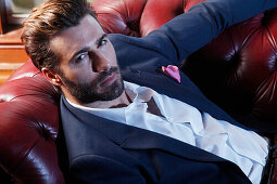 Young man with a beard in a jacket and white shirt lying on a leather sofa