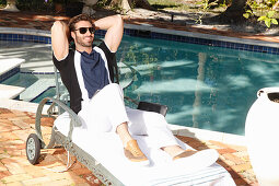 Young man with a beard in a polo shirt and white pants is sitting on a lounger