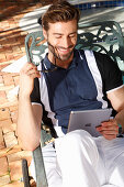 Junger Mann mit Bart in Polo-Shirt und weißer Hose sitzt auf einer Liege, mit Tablet in der Hand