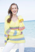 Young, blond woman in yellow jumper with coloured stripes and white trousers at the seaside