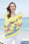 Young, blond woman in yellow jumper with coloured stripes and white trousers at the seaside