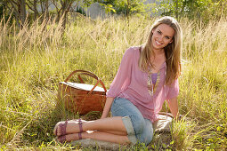 Blonde Frau in rosa Bluse und Jeans-Caprihose mit Picknickkorb auf der Wiese