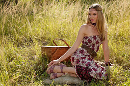 Blonde woman in off-the-shoulder summer dress with picnic basket on the meadow