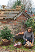 Frau an Feuerstelle im Garten mit kleinem Tannenbäumchen, Picknickkorb, Äpfel auf Gartenbank, vor altem Backhaus